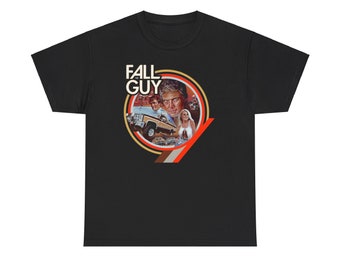 Pode incluir: T-shirt preto com um gráfico circular com o texto "FALL GUY". O gráfico inclui imagens de um homem, uma mulher, um caminhão e outros veículos. O design tem um estilo retrô com detalhes em marrom, laranja e branco.