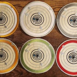 Może przedstawiać: Sześć haftowanych naszywek z białym tłem i czarno-białym wzorem układu słonecznego z planetami krążącymi wokół gwiazdy. Naszywki są okrągłe i mają różne kolory obramowania: złoty, biały, szary, pomarańczowy, zielony i czerwony.