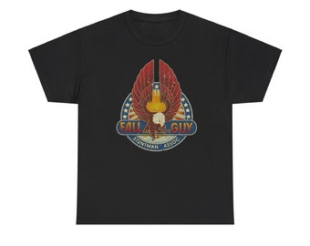 Pode incluir: Camiseta preta com um gráfico desgastado. O gráfico inclui uma águia estilizada com asas vermelhas e amarelas, um círculo azul e branco salpicado de estrelas e o texto "FALL GUY STUNTMAN ASSOC."