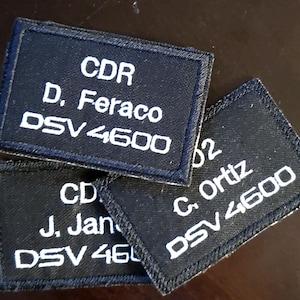 May include: Three black rectangular patches with white text. The patches display names, ranks, and numbers. The top patch reads "CDR D. Feraco DSV 4600". The other patches have similar information.
