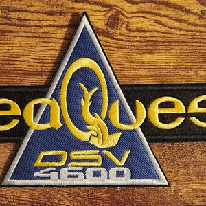 May include: Embroidered patch featuring the text "seaQuest" in gold lettering on a black rectangular background. A blue triangle with gold accents and the text "DSV 4600" is also present. The patch is likely for clothing or accessories.