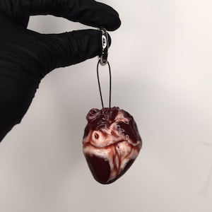 Puede incluir: Colgante en forma de corazón rojo oscuro con vetas blancas y rojas, suspendido de un cordón negro y un anillo plateado. El colgante tiene un acabado brillante y se sostiene sobre un fondo blanco.