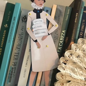 May include: A paper cutout of a woman in a white dress, holding three stacked boxes labeled "GABRIELLE CHANEL." The figure stands in front of a bookshelf filled with books. The woman has a black bow in her hair and wears white shoes.