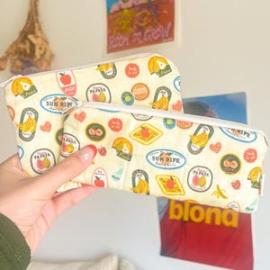 May include: Two cream-colored pencil cases with a repeating pattern of fruit illustrations, including bananas and papayas. The cases have white zippers and are held in a person's hand.