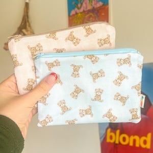 May include: Two small zippered pouches. One is light pink, the other is light blue. Both are covered in a repeating pattern of brown teddy bears. The pouches are held in front of a wall with artwork.