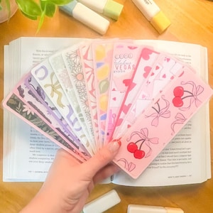 May include: A collection of colorful bookmarks fanned out over an open book. The bookmarks feature various designs, including floral patterns, cherries, and the text "Fabulous Vegas". The bookmarks are held in a hand, with highlighters and other stationery items in the background.