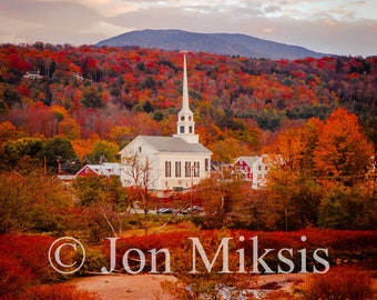 May include: A white church with a steeple stands in a small town surrounded by colorful autumn foliage. The church is nestled at the base of a mountain range with a blue sky and fluffy clouds overhead. The image is captioned "© Jon Miksis".