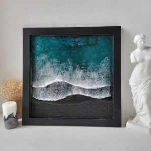 May include: A black framed wall art piece featuring a resin ocean wave crashing on a black sand beach. The wave is white and the water is a teal blue colour.