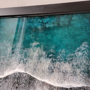 May include: A framed resin art piece depicting a turquoise blue ocean with white foamy waves crashing on the shore.