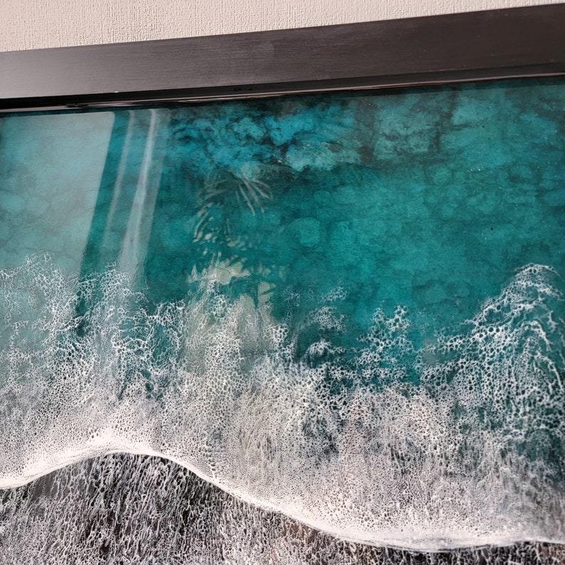 May include: A framed resin art piece depicting a turquoise blue ocean with white foamy waves crashing on the shore.
