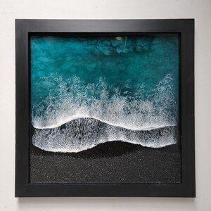 May include: A framed abstract art print depicting a teal blue ocean wave breaking on a black sandy beach. The wave is white and foamy, and the beach has a textured surface.