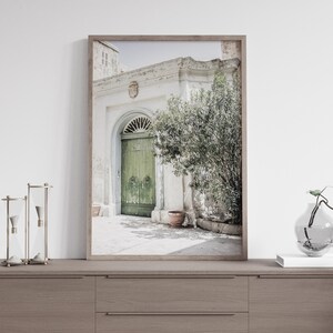 Op de afbeelding: Een ingelijste foto van een groene houten deur met een witte boog en een plant in een pot ervoor. Het gebouw is wit en verweerd. De foto is gemaakt op een zonnige dag buiten.