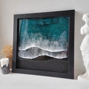 May include: A framed resin art piece depicting a teal ocean with white foamy waves crashing on a black sandy beach.