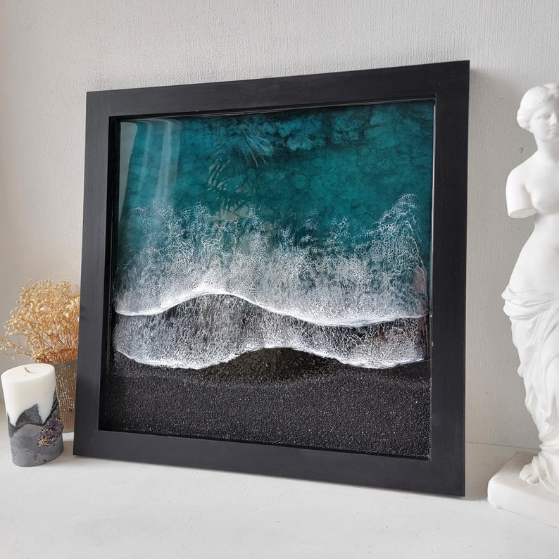 May include: A framed resin art piece depicting a teal ocean with white foamy waves crashing on a black sandy beach.