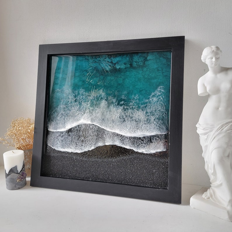 May include: A framed resin art piece depicting a turquoise ocean with white foamy waves crashing on a black sandy beach.