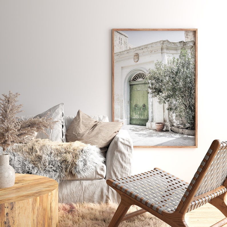 Op de afbeelding: Een woonkamer interieur met een witte bank, een houten stoel, een houten salontafel en een ingelijste foto van een groene deur met een wit gebouw erachter.