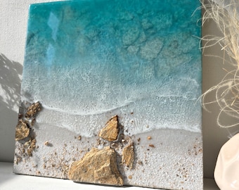 Könnte beinhalten: Quadratisches Kunstwerk aus Kunstharz, das eine Strandszene darstellt. Das Kunstwerk zeigt einen Farbverlauf von Türkis zu Weiß, der das Meer und die Wellen darstellt. Braune Felsen und Sand sind am unteren Rand eingebettet und verleihen der Küstenlandschaft Textur.