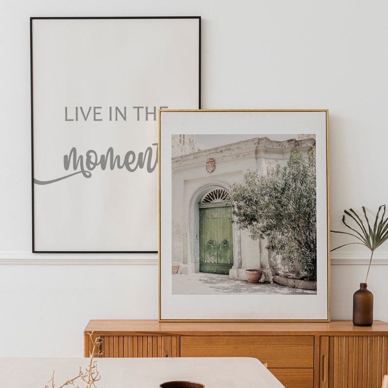 Op de afbeelding: Een ingelijste print van een gebouw met een groene deur en een boom ervoor. De print is in een gouden lijst en heeft een witte passe-partout. De tekst "Live in the moment" is gedrukt op een zwart ingelijste print achter de print van het gebouw.