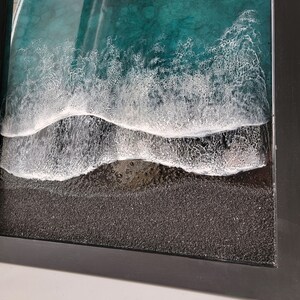 May include: A framed resin art piece depicting a close-up of ocean waves crashing on a black sand beach. The waves are white and foamy, and the sand is textured.