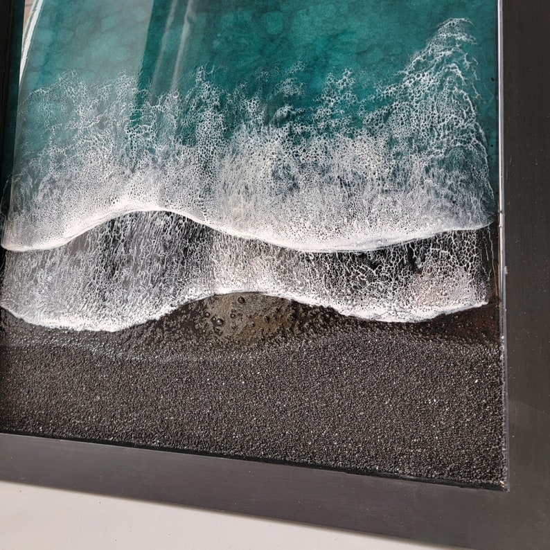 May include: A framed resin art piece depicting a close-up of ocean waves crashing on a black sand beach. The waves are white and foamy, and the sand is textured.