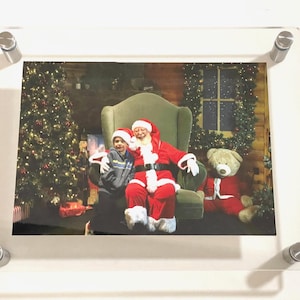 May include: A framed photo of Santa Claus with a child sitting on his lap. Santa is wearing a red suit with white trim and a white beard. The child is wearing a blue and white striped shirt. There is a Christmas tree in the background.