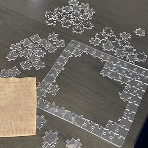 May include: A clear acrylic jigsaw puzzle with a large rectangular frame and smaller pieces scattered around it. The puzzle is on a dark wood surface with a brown fabric bag in the lower left corner.