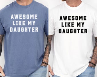 May include: Two t-shirts, one blue and one white, each with the text "AWESOME LIKE MY DAUGHTER" in bold, block letters. The blue shirt is paired with blue jeans, and the white shirt is paired with blue jeans. The bottom of the image has the text "BLUE JEAN" and "WHITE".