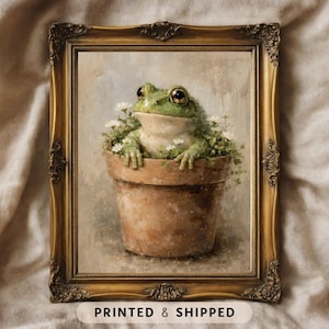 Puede incluir: Impresión artística enmarcada que presenta una rana verde en una maceta marrón con flores blancas. La obra de arte tiene una estética vintage, con un marco dorado ornamentado. El texto "PRINTED & SHIPPED" está en la parte inferior.