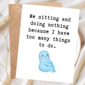 May include: A white greeting card with the text "Me sitting and doing nothing because I have too many things to do." A light blue cartoon character is at the bottom. The card is inside a brown envelope.
