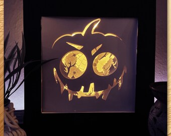 May include: A Halloween-themed, illuminated art piece featuring a carved pumpkin face design. The artwork is set within a black frame, with the pumpkin's eyes and mouth glowing with a warm, golden light. The overall effect is a spooky and festive decoration.