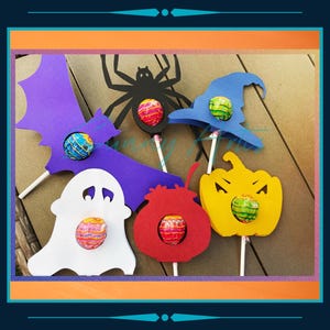 May include: Halloween-themed lollipop decorations. The image features a purple bat, a white ghost, a red cauldron, a yellow pumpkin, and a blue witch hat, each holding a lollipop. A black spider is also present. The decorations are on a brown surface.