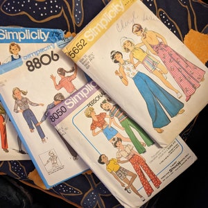 May include: Four vintage sewing patterns for women's clothing. The patterns are from Simplicity and feature different styles of pants and tops. The patterns are in various colors, including blue, pink, and yellow.