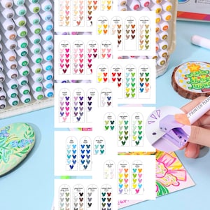 May include: A collection of colorful art markers with a clear plastic case. Color swatches are displayed on small cards, showcasing a wide range of hues, including skin tones, pinks, reds, greens, blues, and browns. A hand holds a marker, demonstrating its use.
