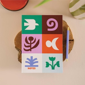 Può includere: Un quaderno colorato con un design geometrico sulla copertina. La copertina presenta uno sfondo verde, marrone, viola, arancione e blu con forme bianche, marroni e verdi. Il quaderno dice "NOTES" in basso.