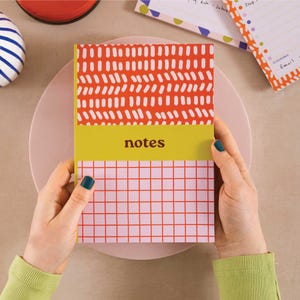 May include: A yellow, orange and pink notebook with a grid pattern on the bottom half and a geometric pattern on the top half. The notebook says "notes" in bold, yellow letters.