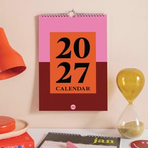 Könnte beinhalten: Ein Wandkalender für das Jahr 2027 mit einem auffälligen Grafikdesign, bestehend aus einem rosafarbenen oberen Bereich, einem dunkelroten unteren Bereich und einem zentralen orangefarbenen Rechteck mit dem schwarzen Text 20 27 CALENDAR.