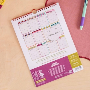 May include: A spiral-bound wall calendar with a white background featuring twelve monthly grid sections in various colorful patterns. The bottom section is purple with white text including the brand name Good Tuesday and recycling icons.