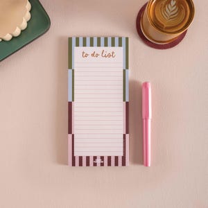May include: A patterned notepad with the words "to do list" in a handwritten font. A pink pen lies next to the notepad. The notepad features a pink, brown, blue, and green colour scheme. The notepad is lined.