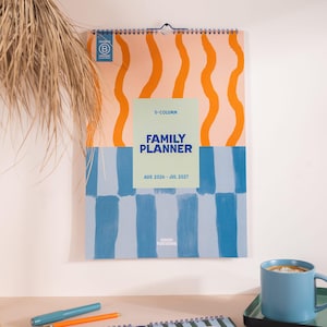 May include: A family planner calendar with a spiral binding. The top section has orange wavy lines on a peach background, and the bottom has a blue and white checkered pattern. The calendar is labelled "Family Planner" and covers August 2026 to July 2027.