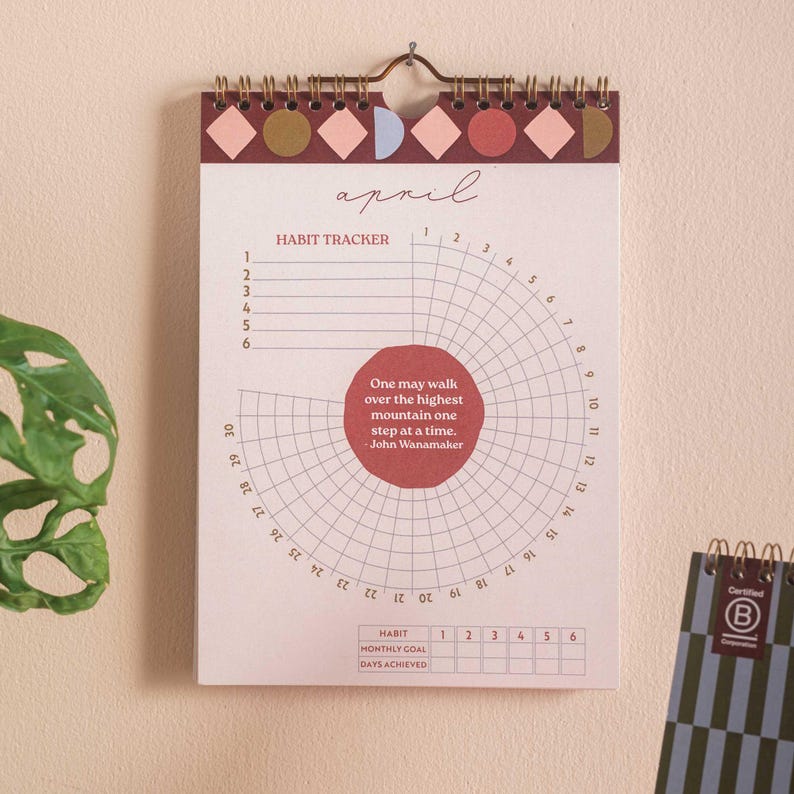 May include: A wall calendar for April with a habit tracker. The top border has geometric shapes in pink, brown, and blue. The calendar includes a quote by John Wanamaker: "One may walk over the highest mountain one step at a time."
