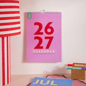 May include: A pink wall calendar with the numbers "26" and "27" in red. The word "CALENDAR" is below the numbers. The calendar is hanging on a white wall. A red and white striped lamp is on the left.