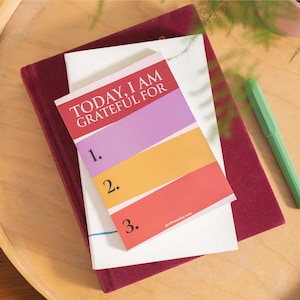Puede incluir: Un diario de gratitud colorido con el texto "TODAY, I AM GRATEFUL FOR" y líneas numeradas para escribir. El diario tiene una cubierta a rayas de color morado, amarillo y rojo con el sitio web "gratitudesaday.com" impreso en la parte inferior.