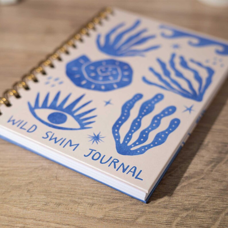 May include: A cream-colored spiral bound journal with a blue and white design. The cover features a stylized eye, seaweed, and the text "WILD SWIM JOURNAL".