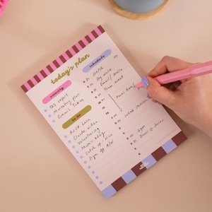 May include: A rectangular daily planner notepad with a striped border in brown, pink, and purple. The page features sections titled today's plan, schedule, priority, and to do, with handwritten tasks and times written in black ink using a pink pen.