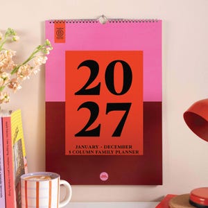 May include: A wall-mounted 2027 calendar with a spiral binding, featuring a pink top section and a dark maroon bottom section. A large orange rectangle in the center displays the year 2027 in black text, with January-December 5 Column Family Planner below.