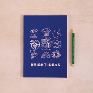 Könnte beinhalten: Ein blaues Notizbuch mit einem rosafarbenen Design aus verschiedenen Illustrationen von Meereslebewesen. Das Notizbuch trägt den Text "BRIGHT IDEAS" auf der Vorderseite.
