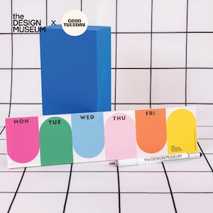 Puede incluir: Un bloc de notas semanal horizontal con cinco secciones coloridas de parte superior redondeada, etiquetadas como MON, TUE, WED, THU y FRI. Un bolígrafo blanco con el texto "the DESIGN MUSEUM" descansa delante. Un bloque rectangular azul está detrás.