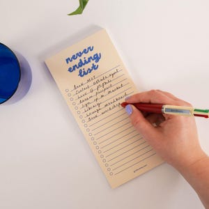 Könnte beinhalten: Ein cremefarbener Notizblock mit dem blauen Text "never ending list" oben, der eine Spalte mit leeren Kreisen und horizontalen Linien für Aufgaben enthält. Ein mehrfarbiger Kugelschreiber mit roten, grünen und weißen Teilen schreibt auf das Papier.