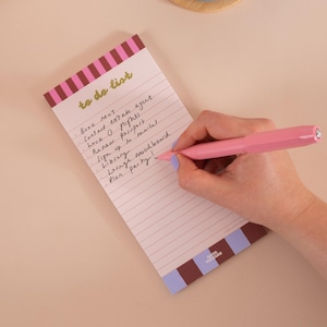 May include: A rectangular to-do list notepad with pink and brown striped borders at the top and bottom. The paper features horizontal lines and the text to do list in gold script. A pink pen is writing on the paper, which contains handwritten notes.