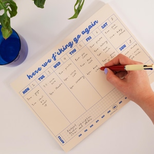 May include: A cream-coloured weekly planner pad with blue text that reads "here we f*cking go again" at the top. The grid features days of the week from Monday to Sunday with handwritten appointments in black ink and a notes section at the bottom.